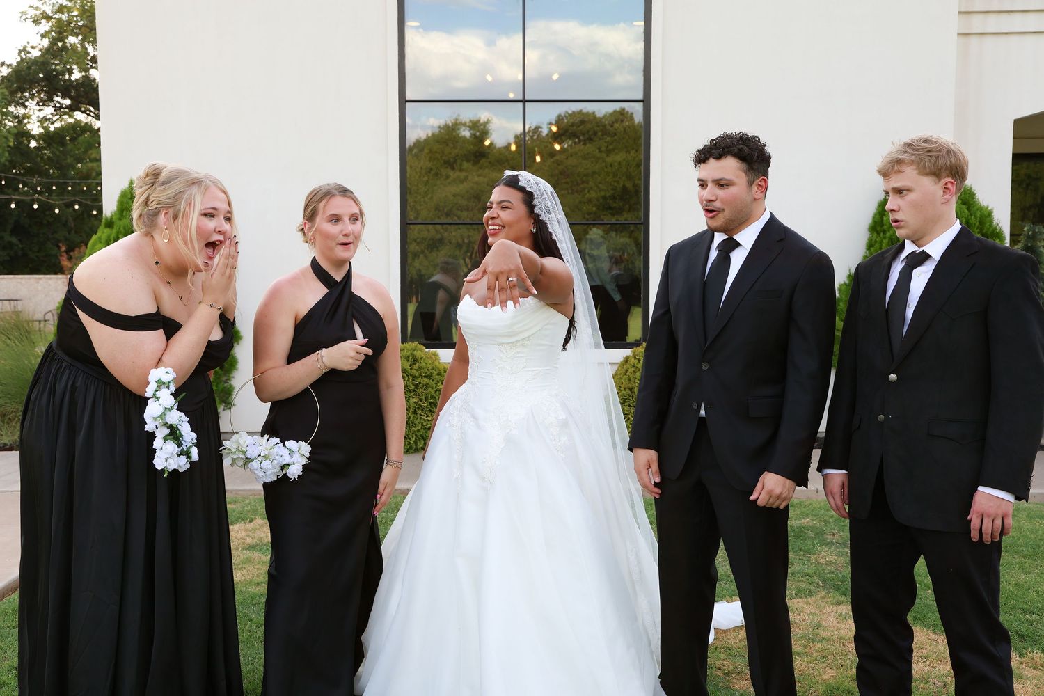 Bride in white ball gown poses with bridesmaids in black dresses and groomsmen in suits at outdoor wedding.