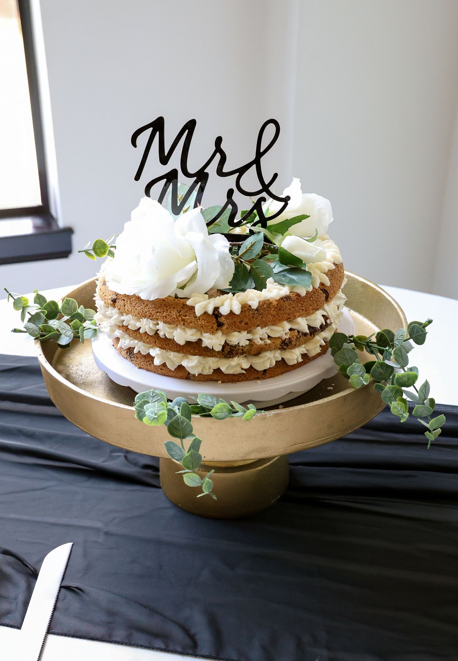 Rustic naked wedding cake with Mr & Mrs topper, white roses, and greenery on a gold cake stand.