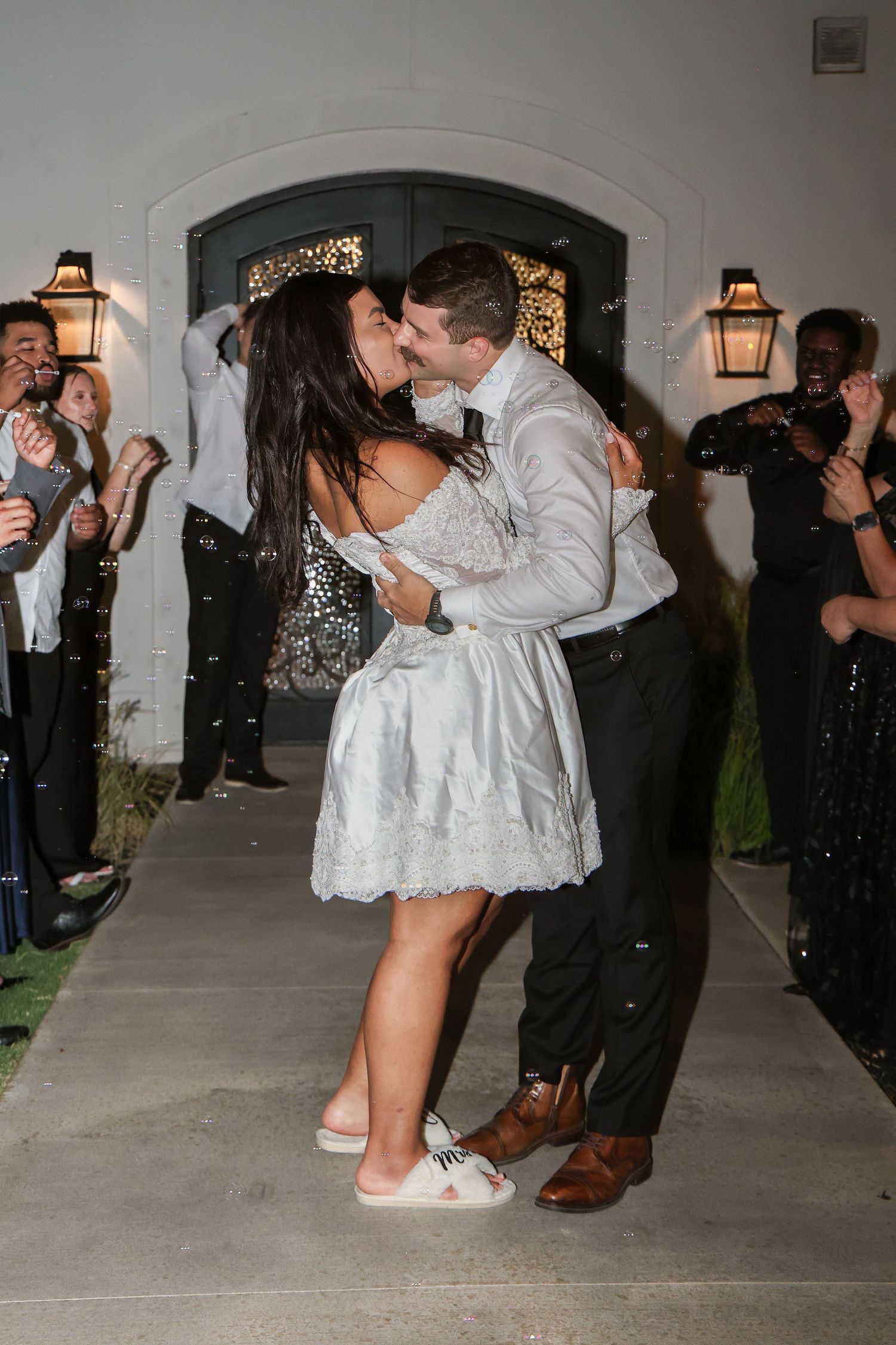 Bride and groom share a kiss during their wedding send-off with guests celebrating around them at night.
