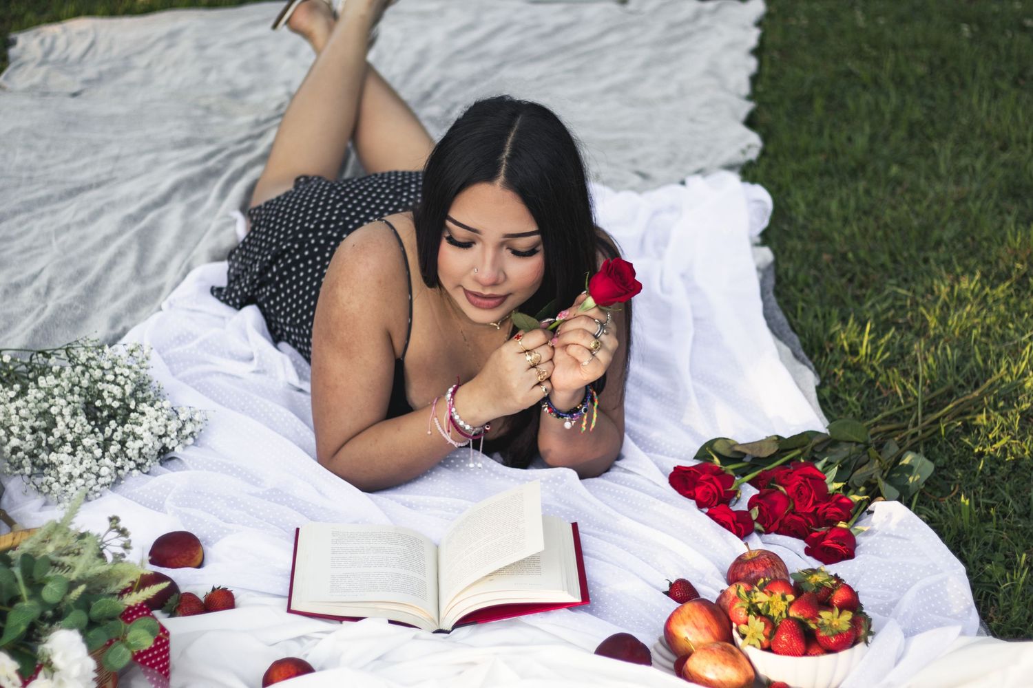 Young woman lying on white blanket outdoors, holding a red rose near an open book, surrounded by fresh strawberries and flowers.