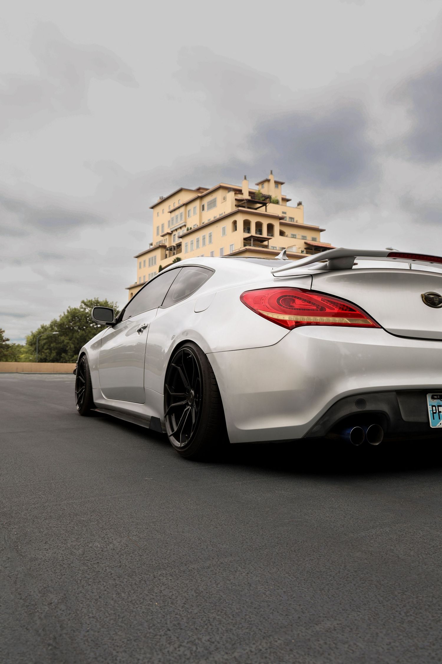 White Infiniti G37 coupe with black wheels and rear spoiler parked on asphalt with a beige mansion in the background.