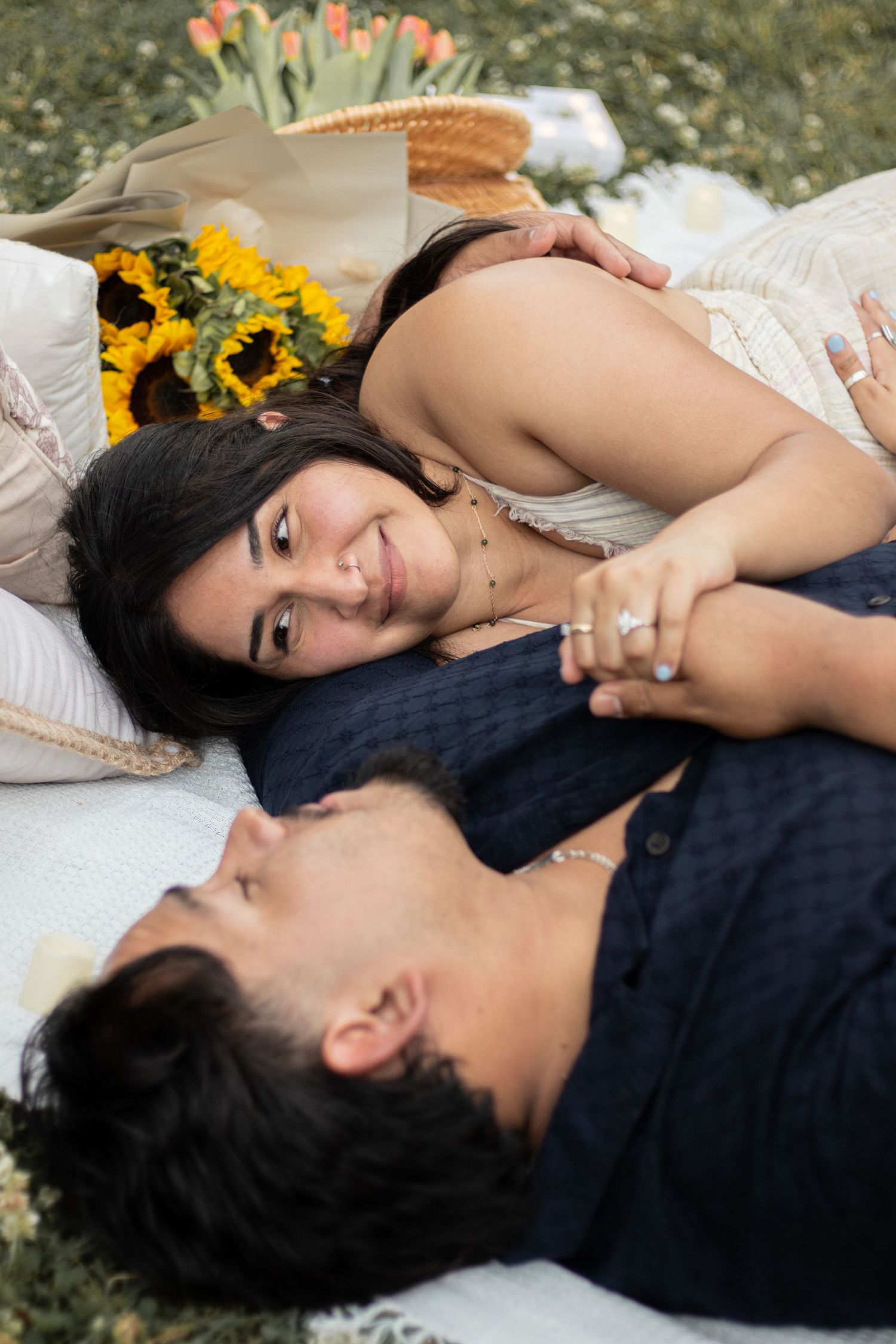 Couple lying on a blanket outdoors, holding hands and smiling, with sunflowers and tulips nearby.