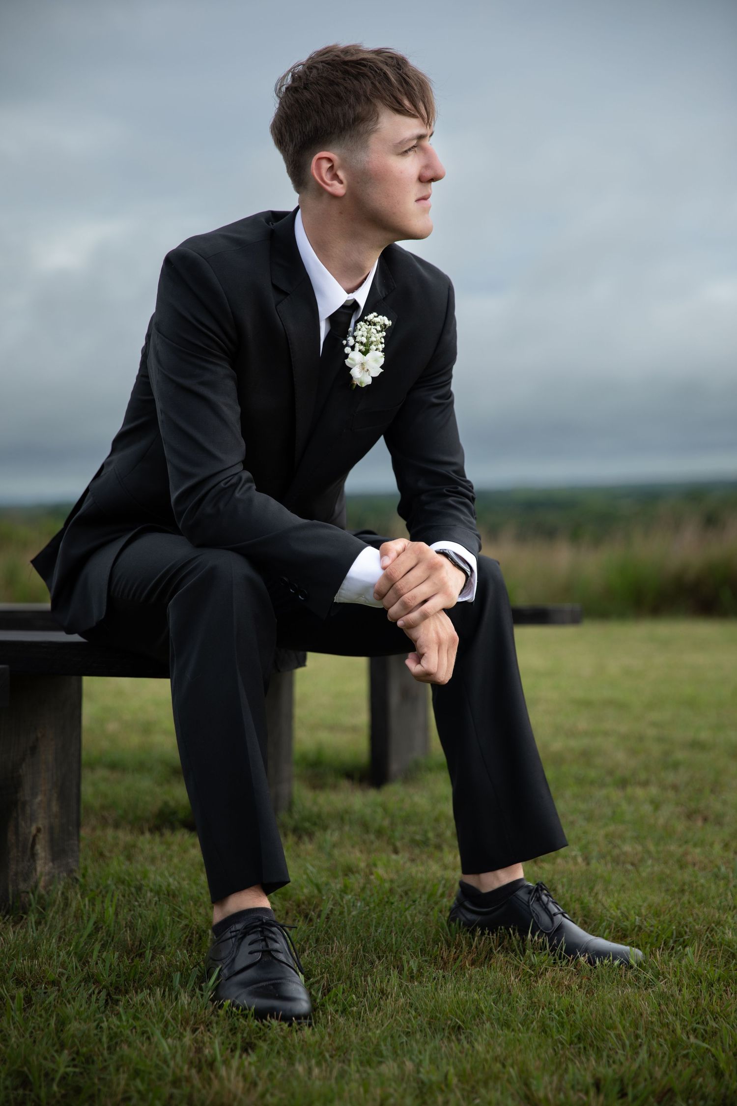 Young man in black suit with boutonniere sits on bench outdoors, gazing thoughtfully against cloudy sky.
