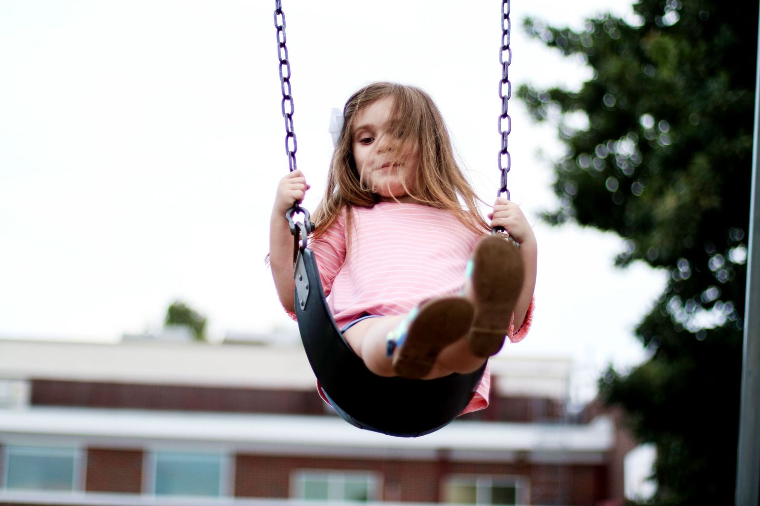 Someone wearing a pink shirt enjoys swinging on a playground swing set against a blurred building background.