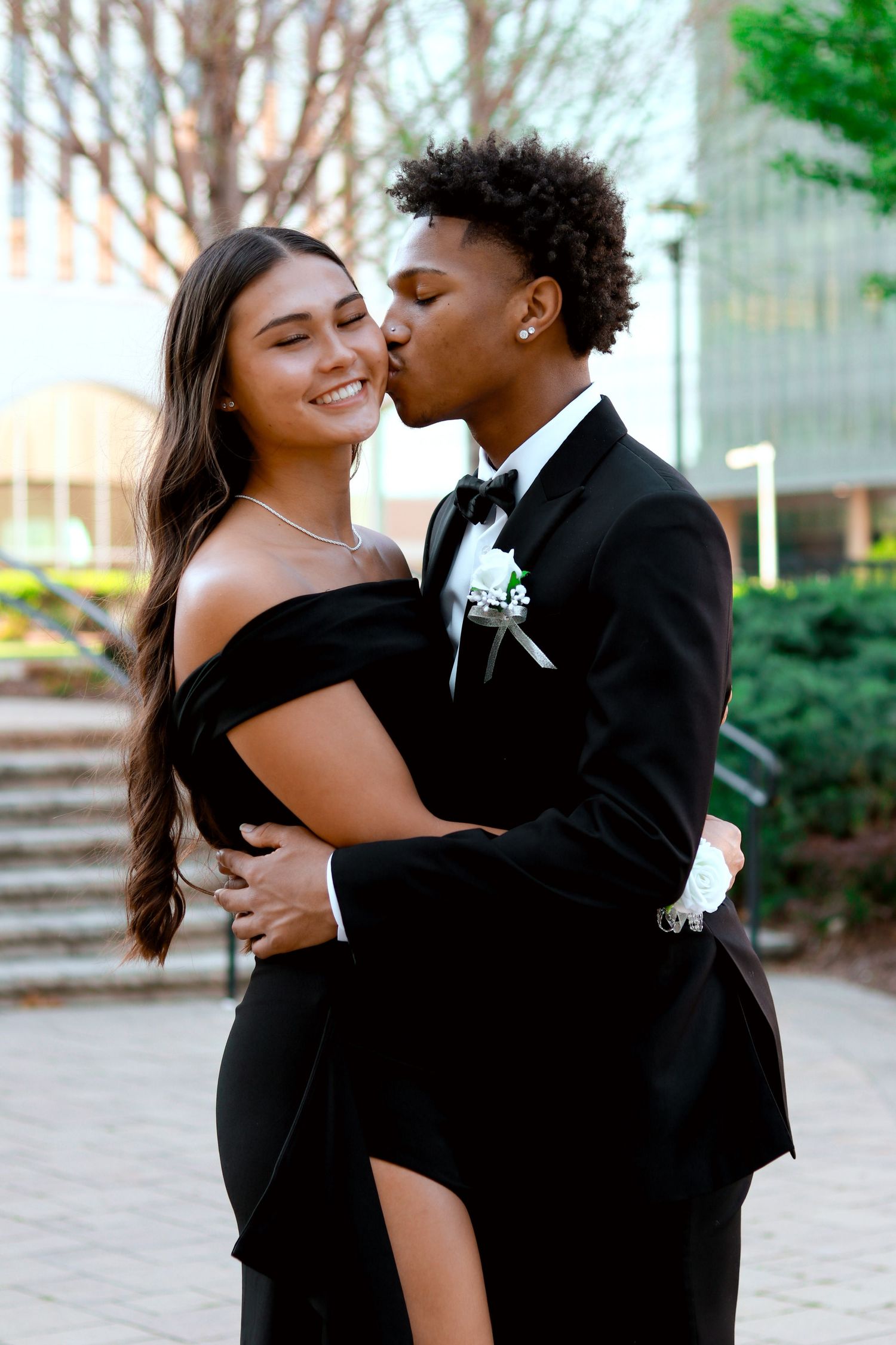 A romantic couple in formal black attire embraces and shares intimate moments during a photo session outdoors.