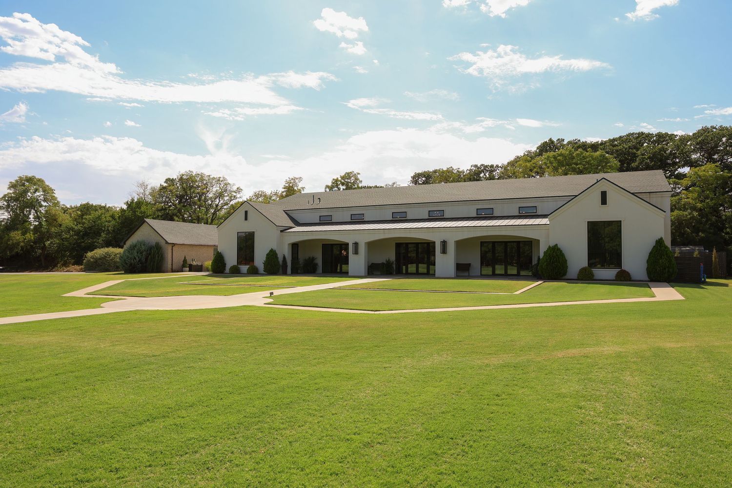Large white farmhouse-style estate with expansive green lawn, covered porch, and surrounded by mature trees on a sunny day.