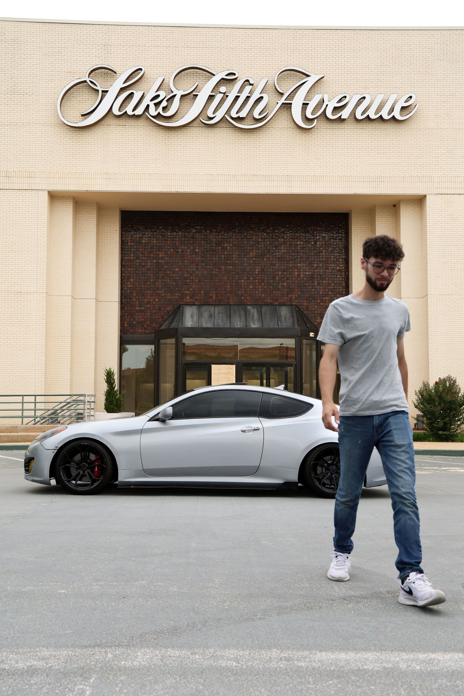 Young man in gray t-shirt and jeans poses before Saks Fifth Avenue with a matte gray sports car.