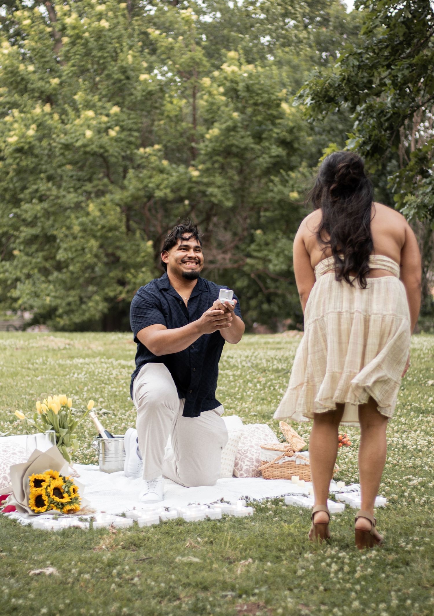 Man on one knee proposing to woman during romantic outdoor picnic surrounded by sunflowers and champagne.