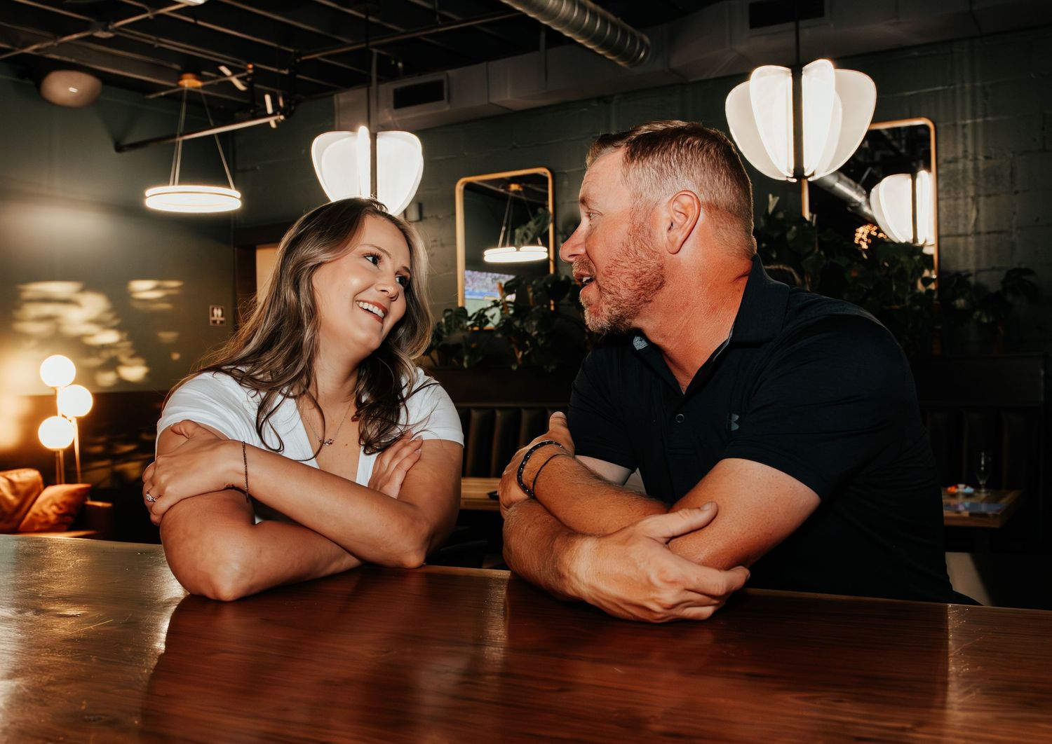 A man and woman smile at each other while leaning on a bar counter in a warmly lit restaurant.