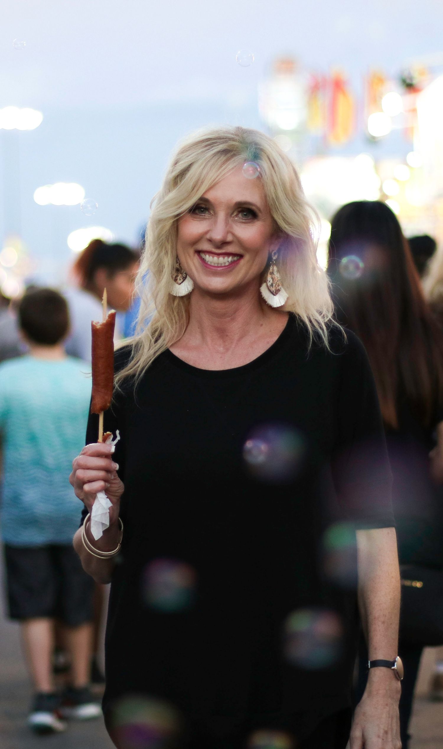 A person in a black outfit smiling eating fair food with bubbles and carnival games in the background.