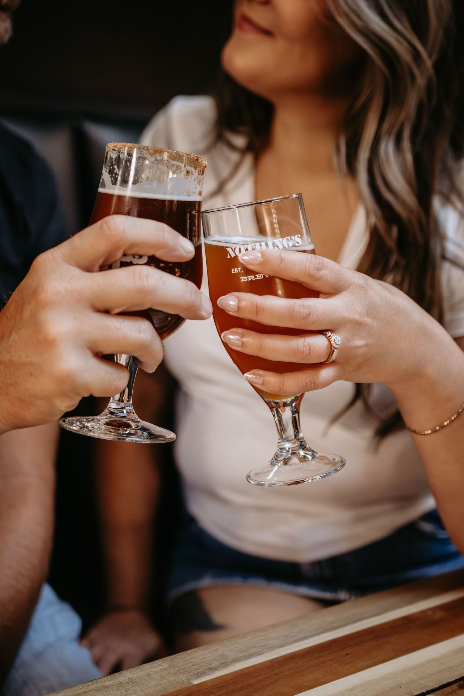 Couple clinking glasses of craft beer in branded pilsner glasses at a brewery, woman wearing engagement ring.