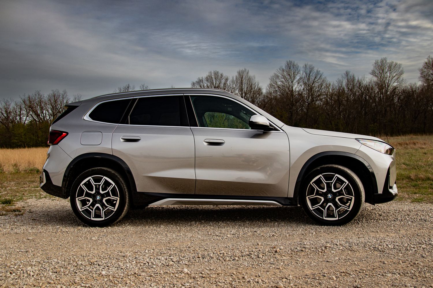 Silver SUV crossover parked on gravel road, side profile view, stylish alloy wheels, overcast sky background.