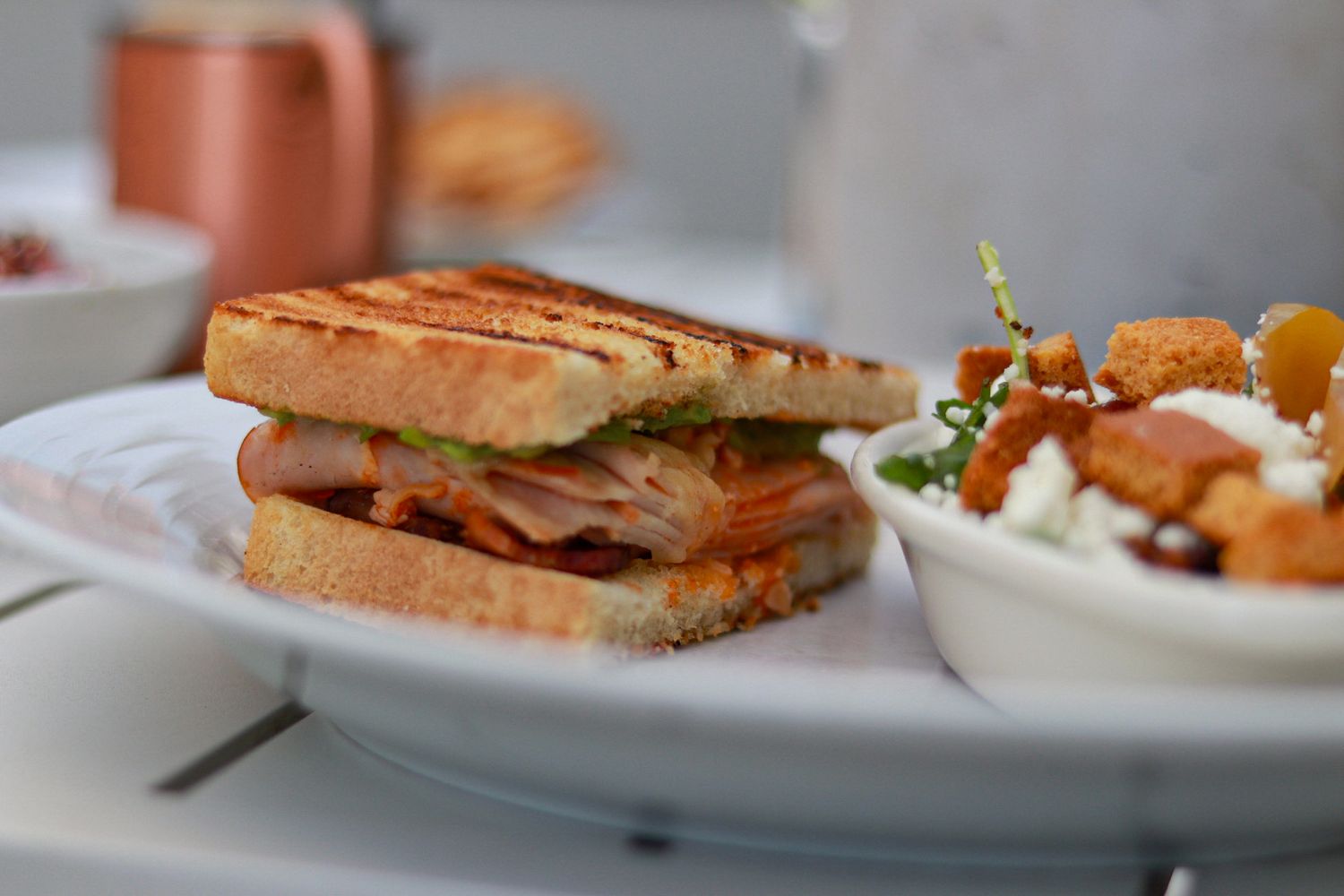 Grilled panini sandwich with turkey and pesto on toasted bread, served with a side salad on a white plate.