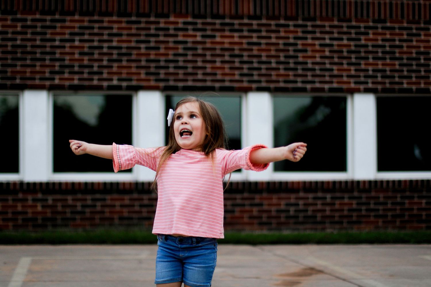 A child in a pink shirt and jeans stands with arms outstretched in front of a brick building.