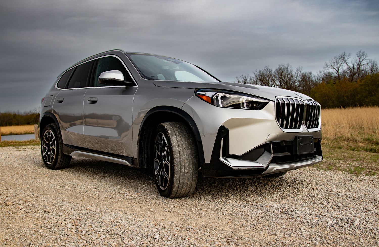Silver BMW X1 SUV parked on gravel with dramatic cloudy sky and bare trees in background.