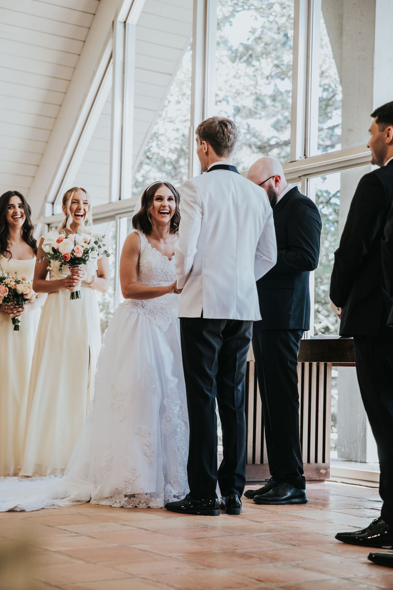 Bride laughing joyfully during indoor wedding ceremony with bridesmaids holding bouquets and groom in white jacket.