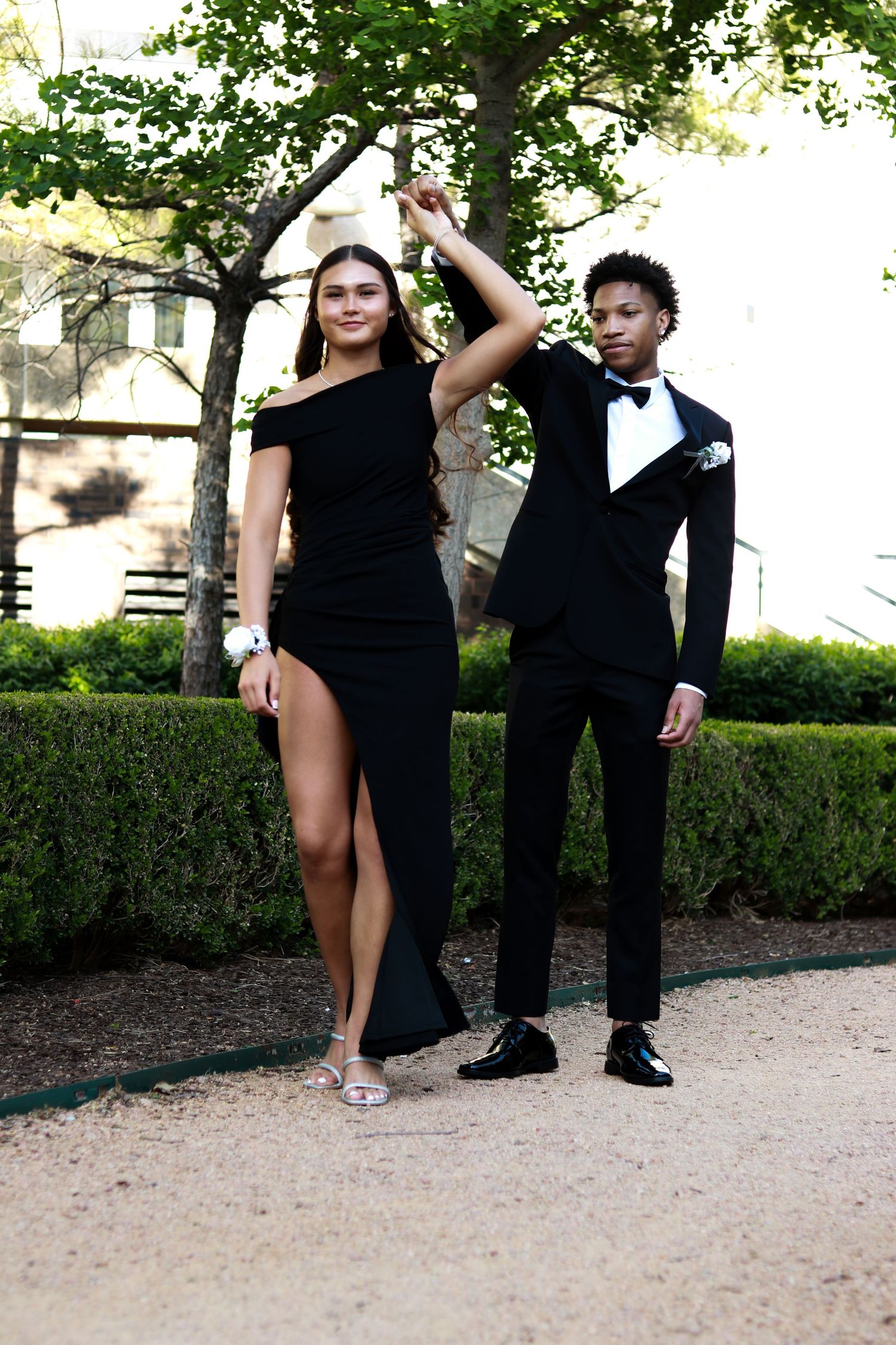 A couple poses for prom photos outdoors, wearing elegant black formal attire on a garden path.