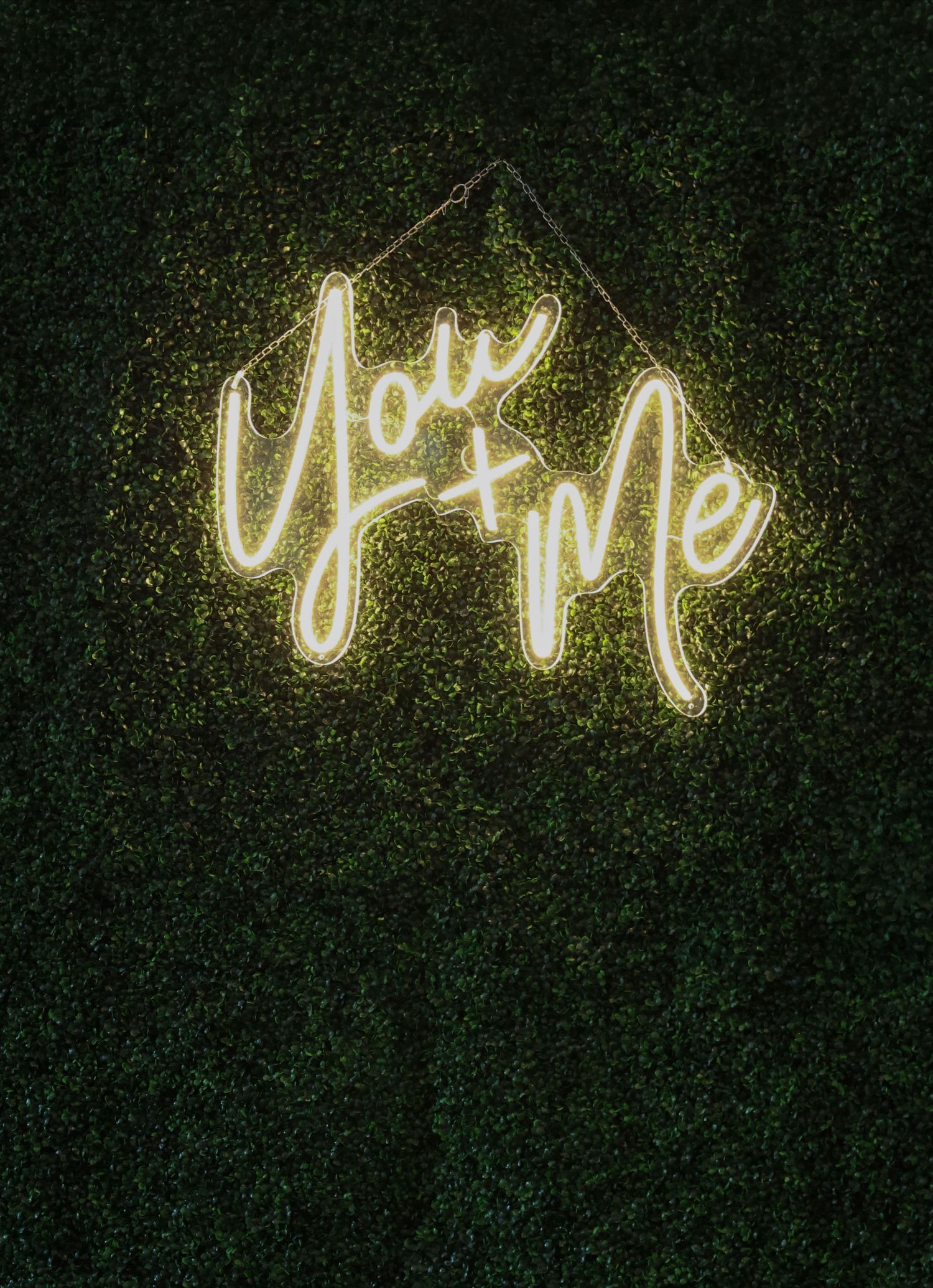 Glowing neon sign reading You + Me in warm white light mounted on a lush green hedge wall backdrop.