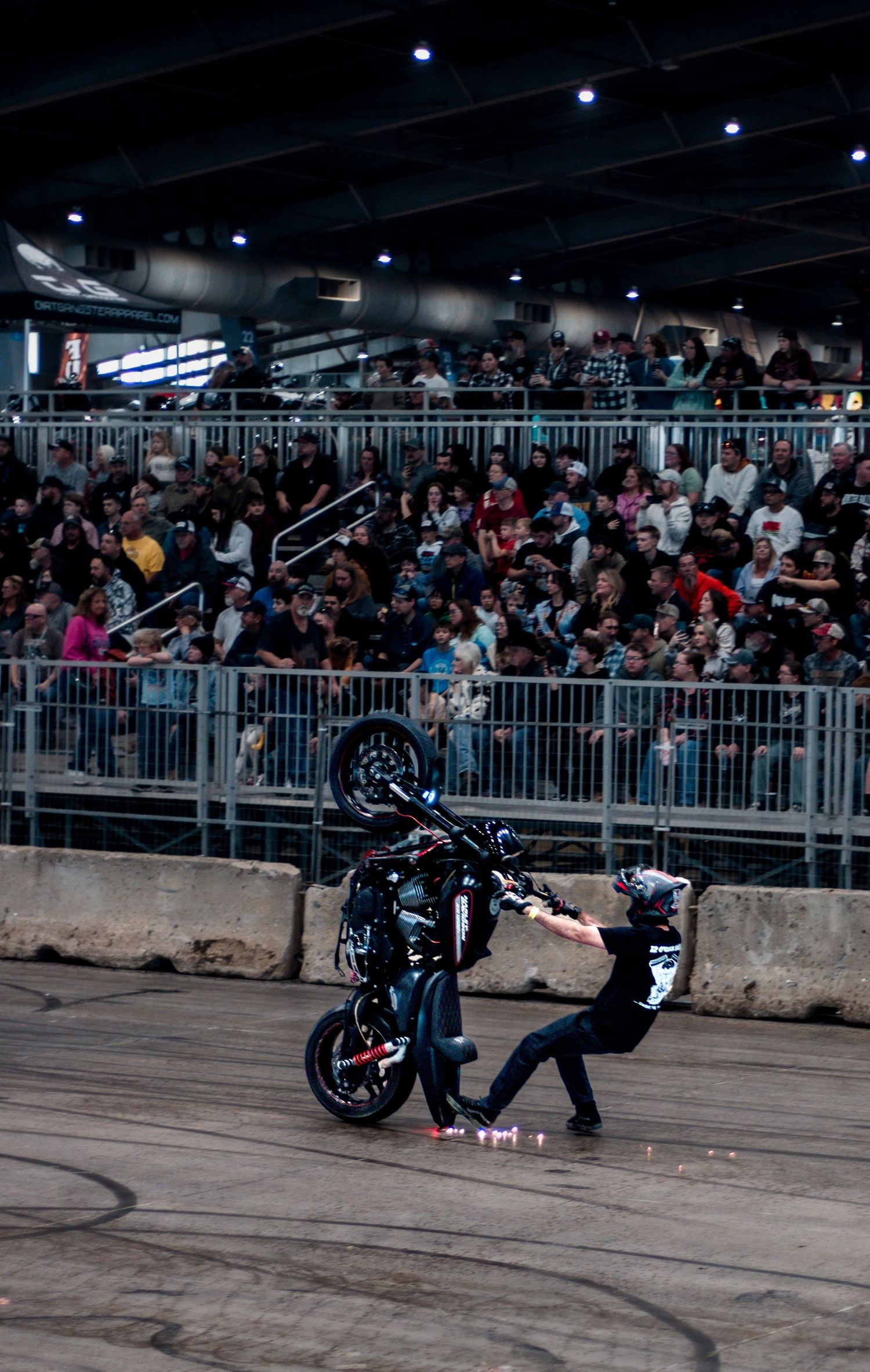 A stunt rider performs a dramatic motorcycle wheelie trick before a large crowd at an indoor arena event.