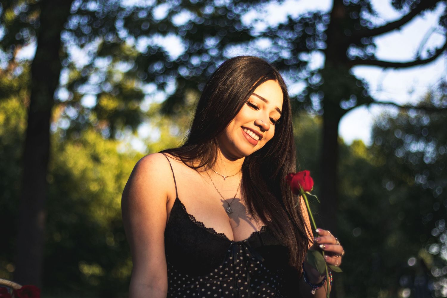Young Asian woman in black dress smiling while holding a red rose outdoors in golden sunlight.