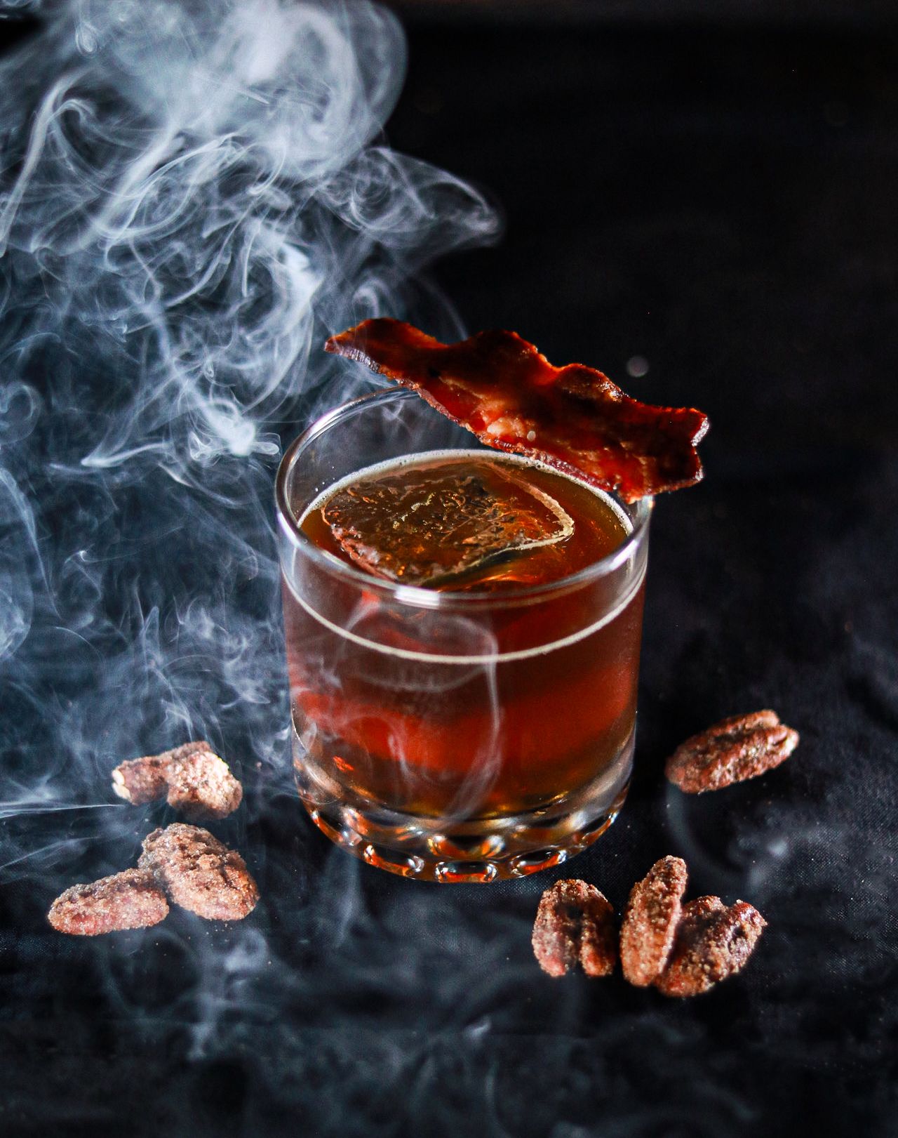 Smoky whiskey cocktail in rocks glass with candied pecans and crispy bacon garnish on dark background.