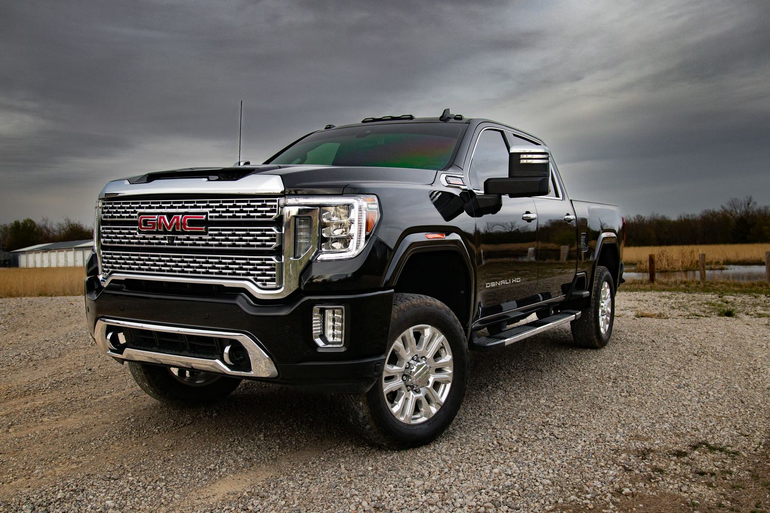 Black GMC Sierra 2500HD Denali pickup truck parked on gravel under dramatic cloudy sky, front three-quarter view.