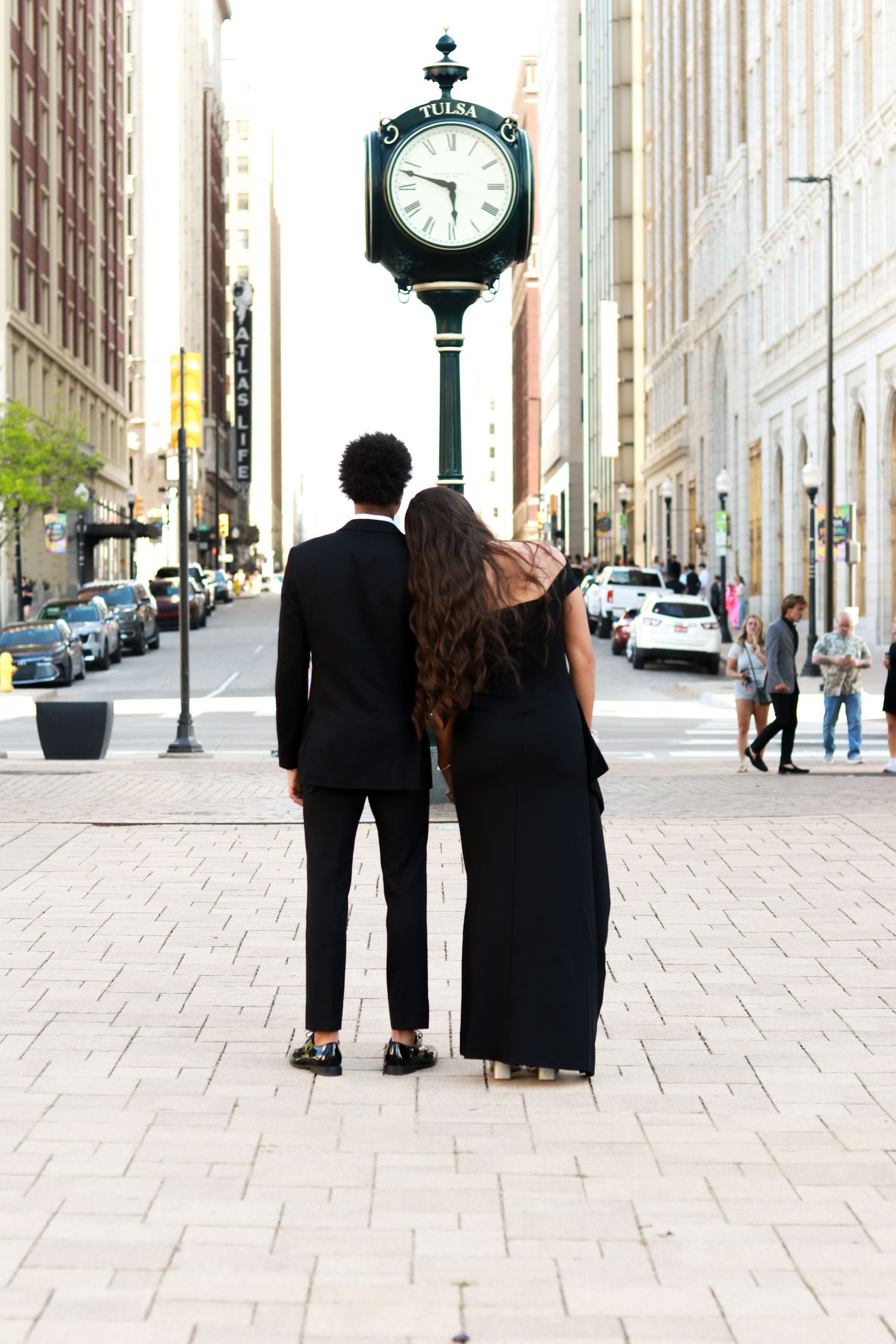 A couple in formal black attire walking away from camera on a stone sidewalk near a decorative street clock.