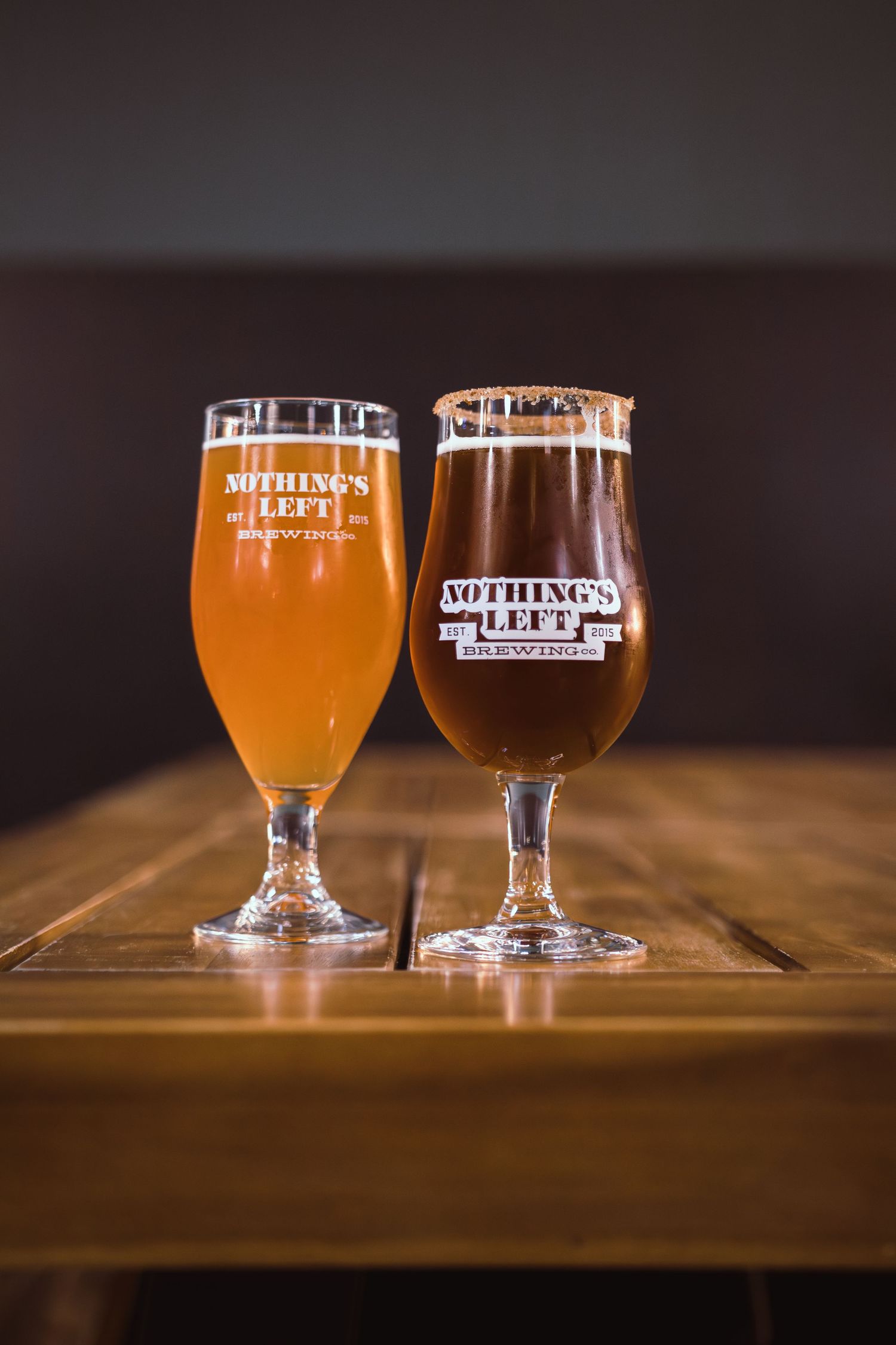 Two craft beers in tulip glasses branded Nothing's Left Brewing Co. on a wooden table.