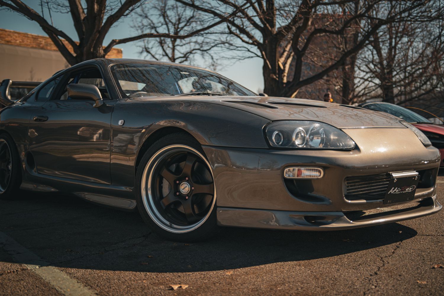 Silver Toyota Supra MK4 parked outdoors, featuring wide body kit, deep dish wheels, and aggressive front bumper styling.