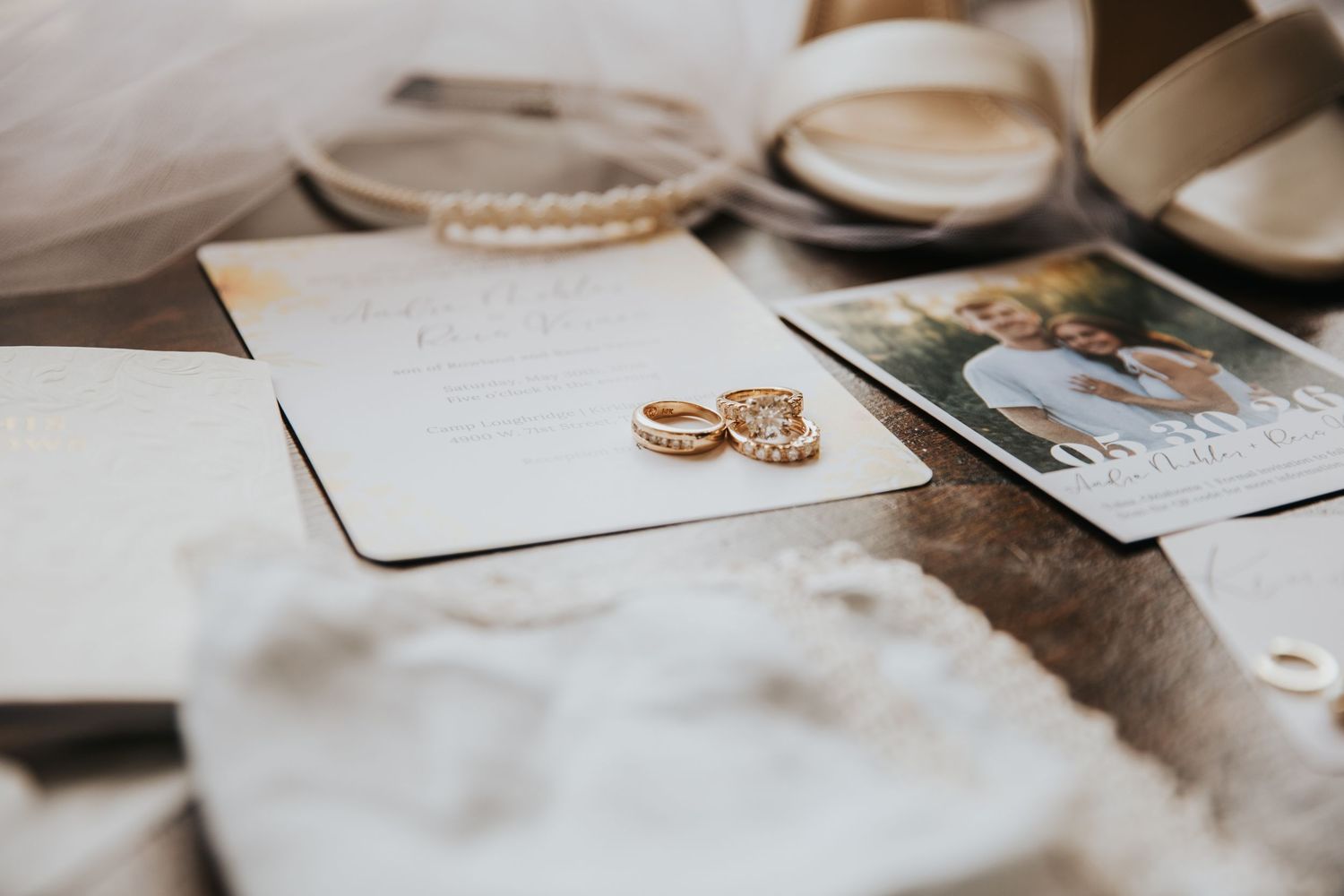 Gold wedding rings resting on elegant wedding invitation cards with a couple's photo and bridal accessories on rustic wood.
