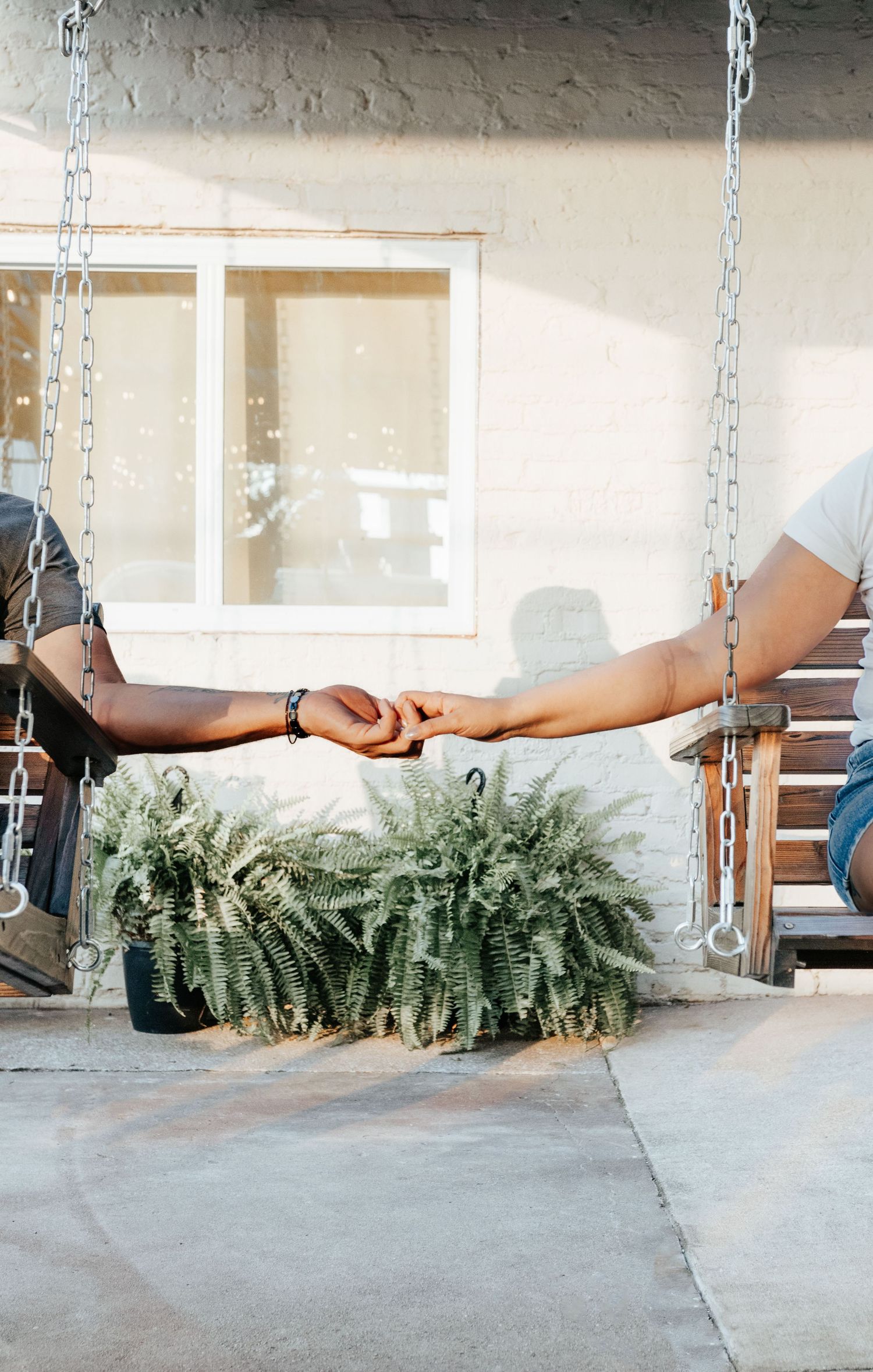 Two people holding hands across porch swings with a fern plant between them near a white building.