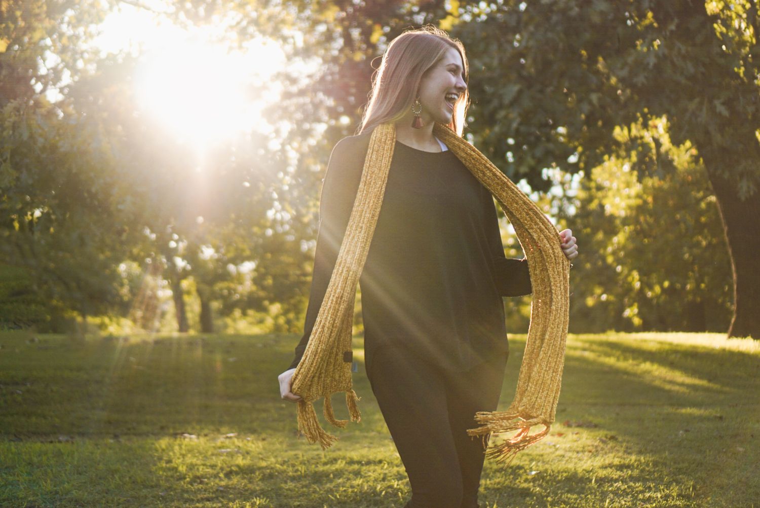 Person wearing black dress and yellow scarf stands in sunlit garden during golden hour.