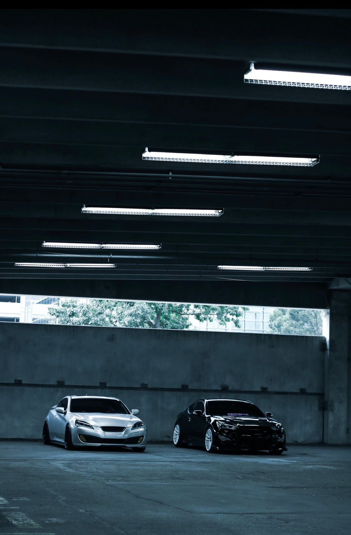 Two lowered sports cars, one white and one black, parked in a dimly lit parking garage with fluorescent lights.