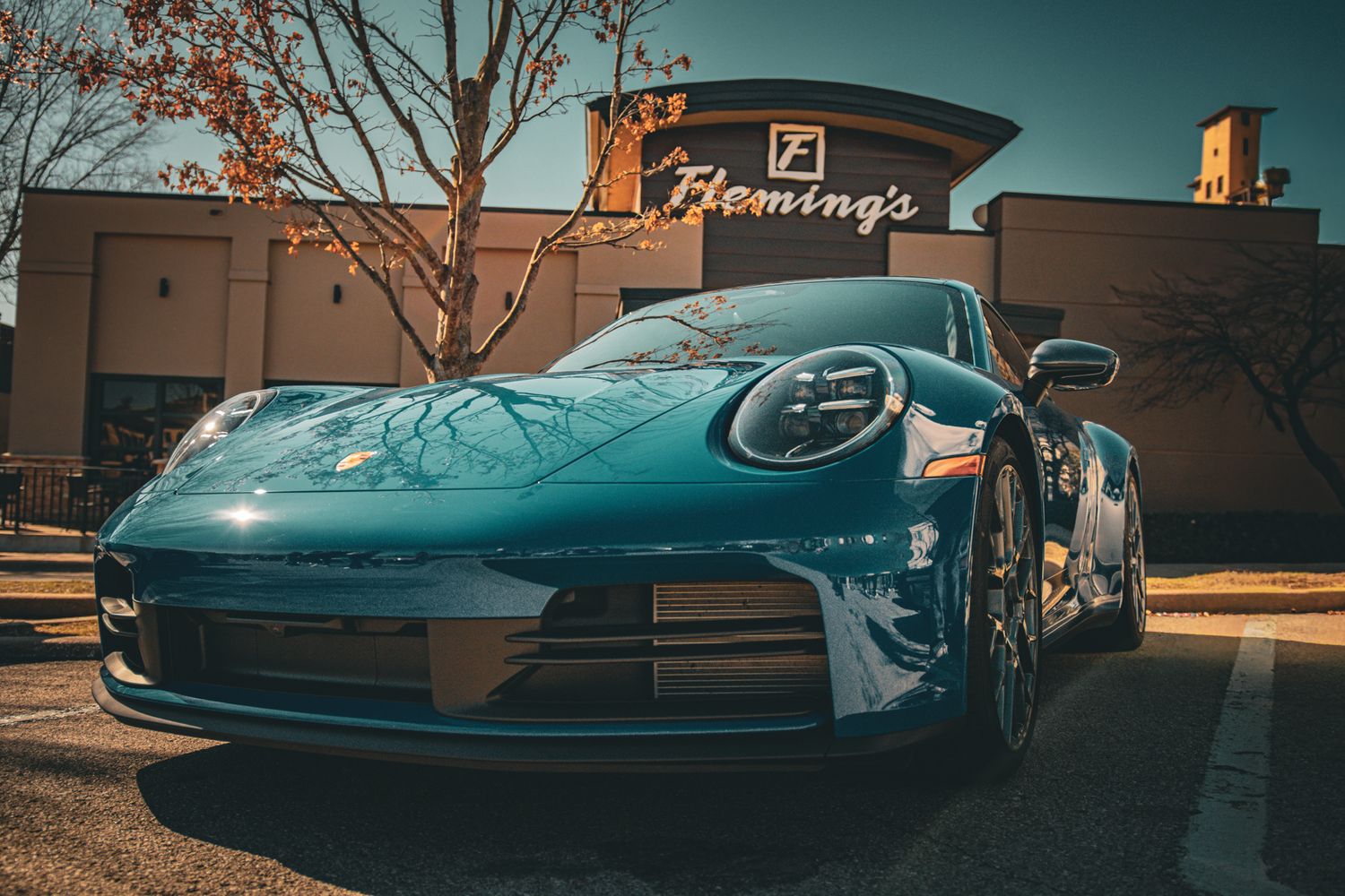 A sleek blue Porsche 911 parked outside Fleming's restaurant on a sunny day with autumn trees in the background.