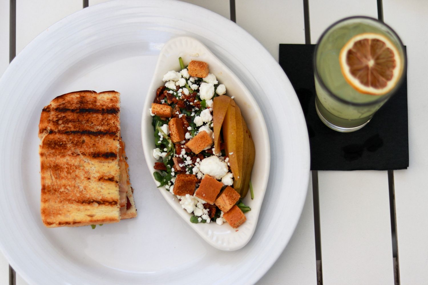 A white plate with a grilled sandwich, veggie taco with feta cheese, and a green drink garnished with lemon.