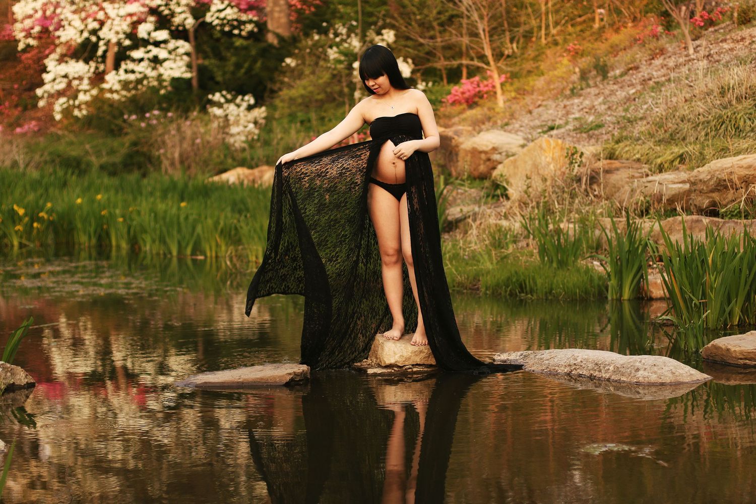 A figure in a black dress stands elegantly by a tranquil pond surrounded by autumn foliage and rocks.