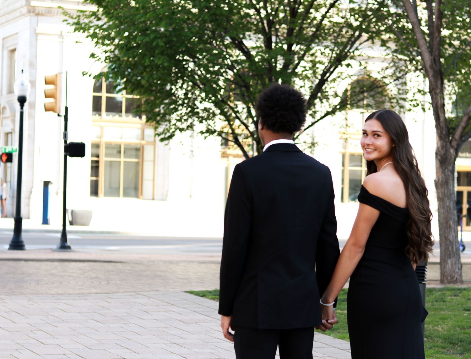 A couple in formal attire walks together on a tree-lined urban sidewalk on a sunny day.