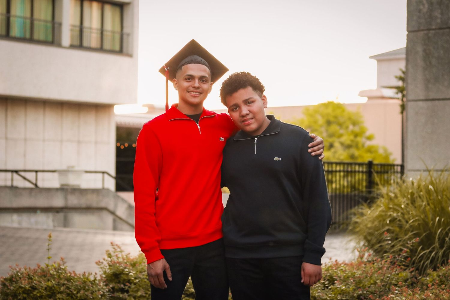 Two people in red and black outfits pose together outdoors near a building during daytime.