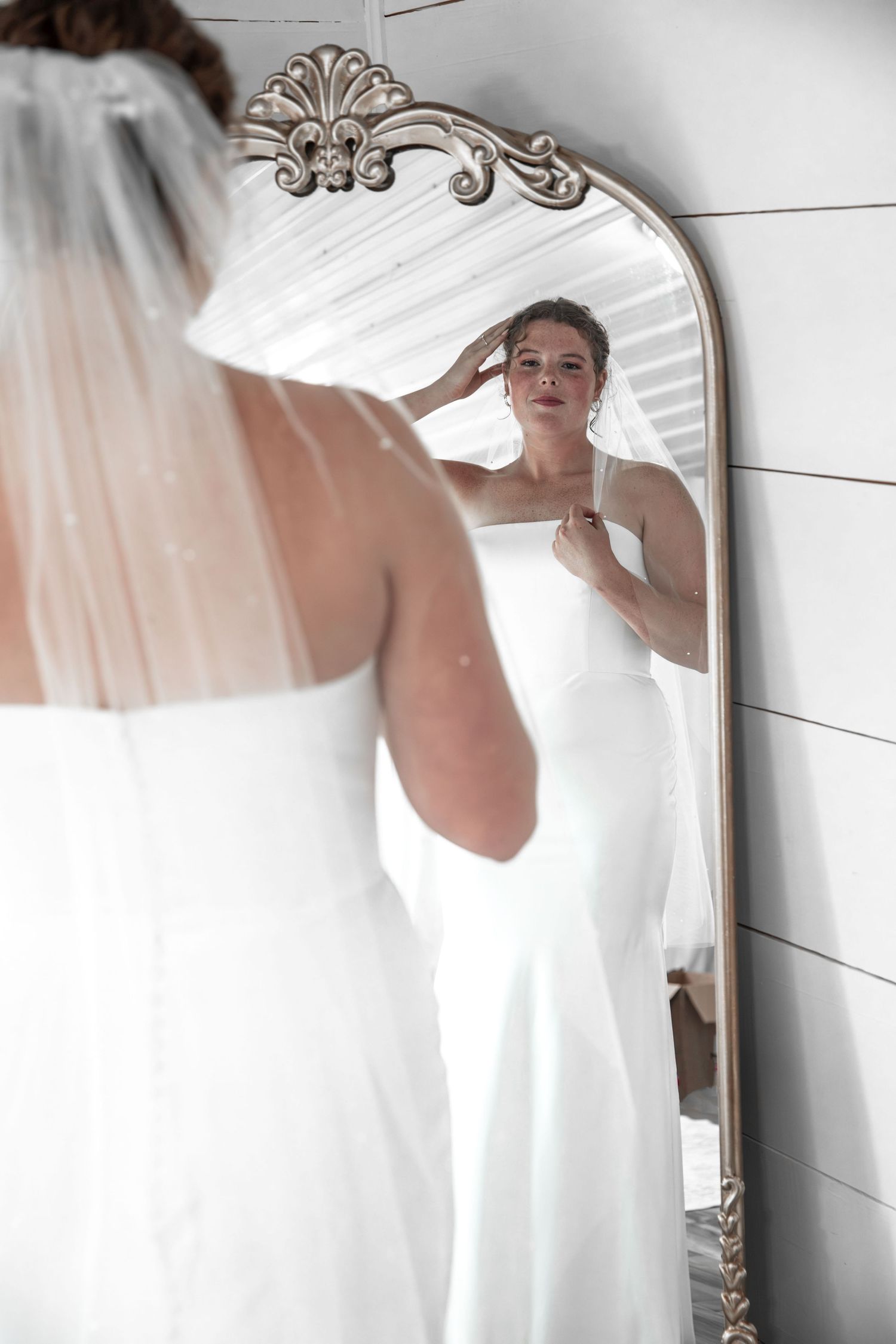 Bride in white gown adjusting dress while viewing reflection in ornate gold-framed mirror.