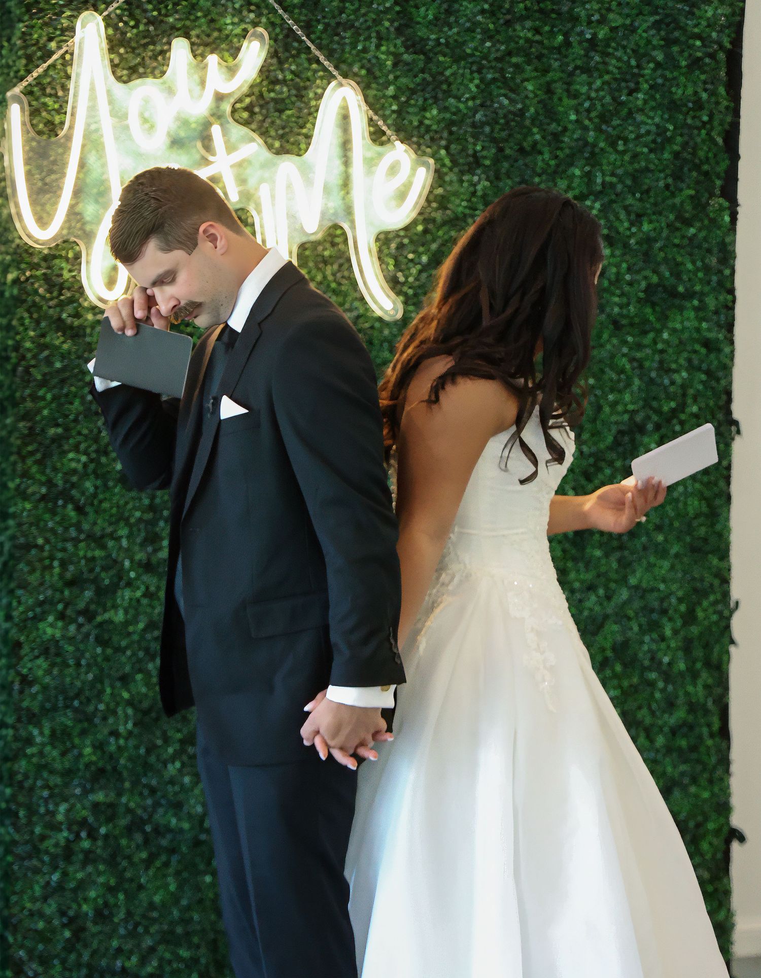 Bride and groom share private vows back-to-back before a green hedge wall with a glowing You & Me neon sign.