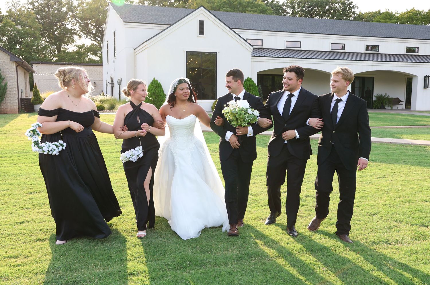 Bride in white gown with bridesmaids in black dresses and groomsmen in suits walking on green lawn outside white venue.