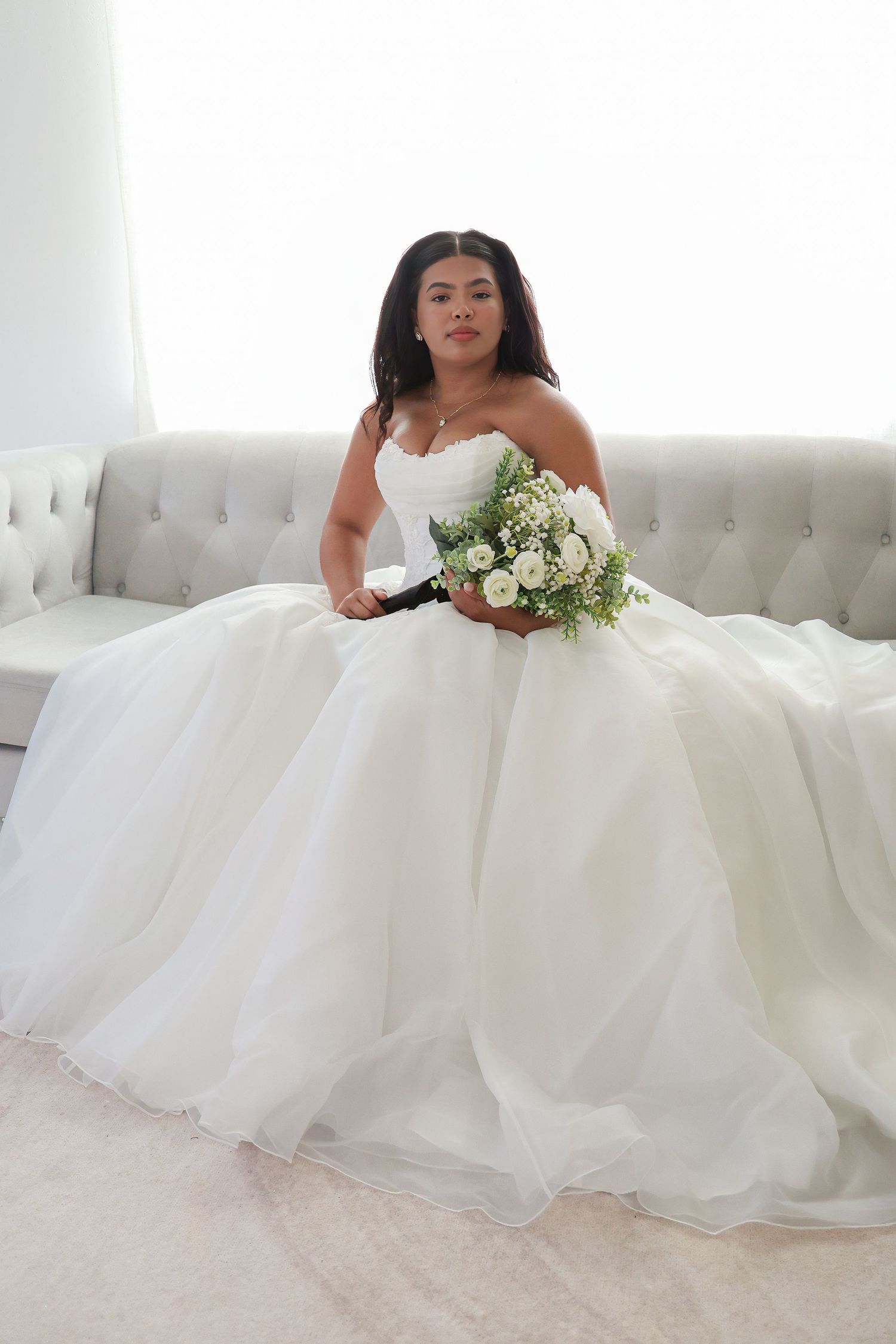 Bride in strapless white ball gown holding white floral bouquet, seated on gray tufted sofa in bright room.