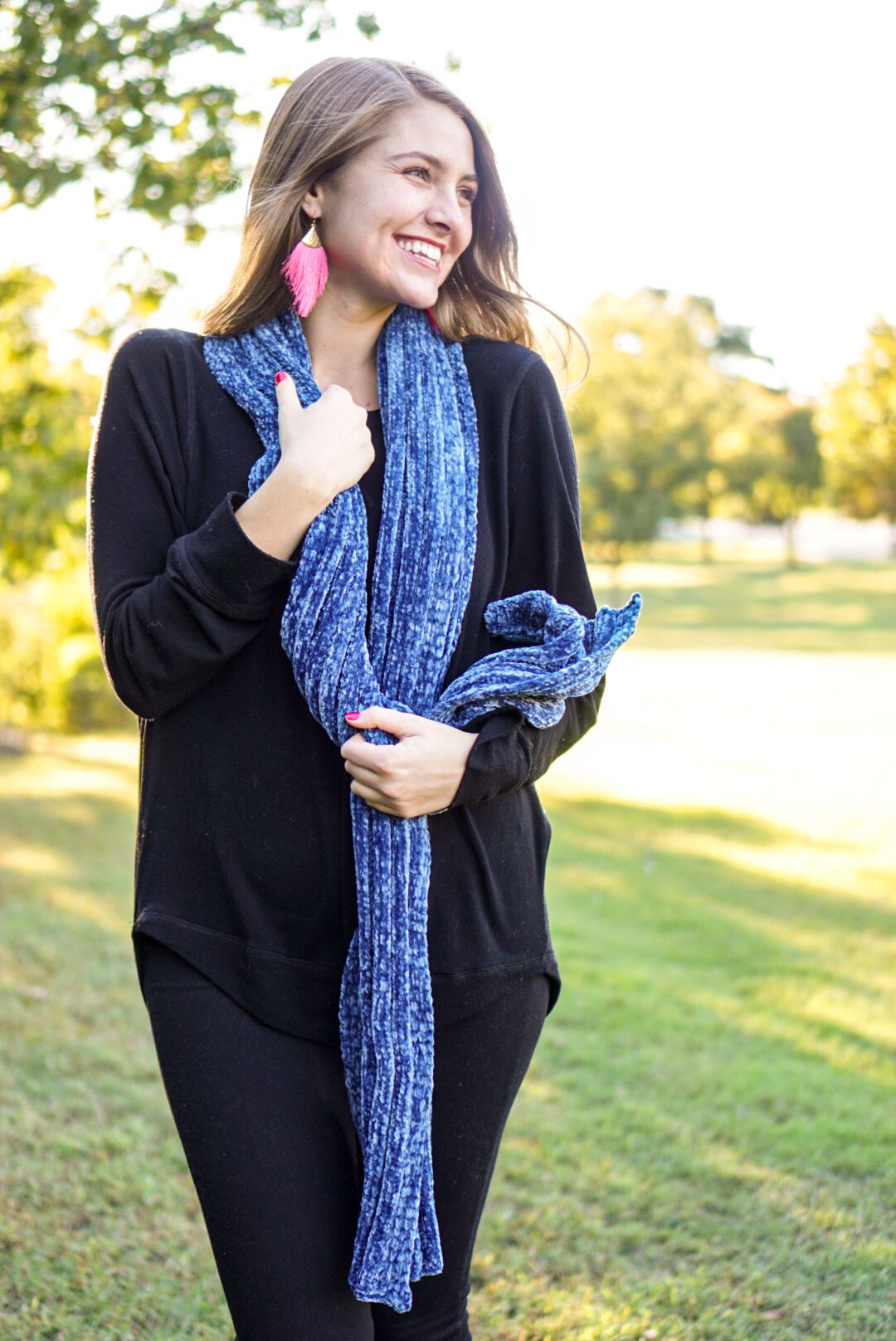 A person wearing a black sweater and blue scarf poses outdoors in natural sunlight with pink statement earrings.