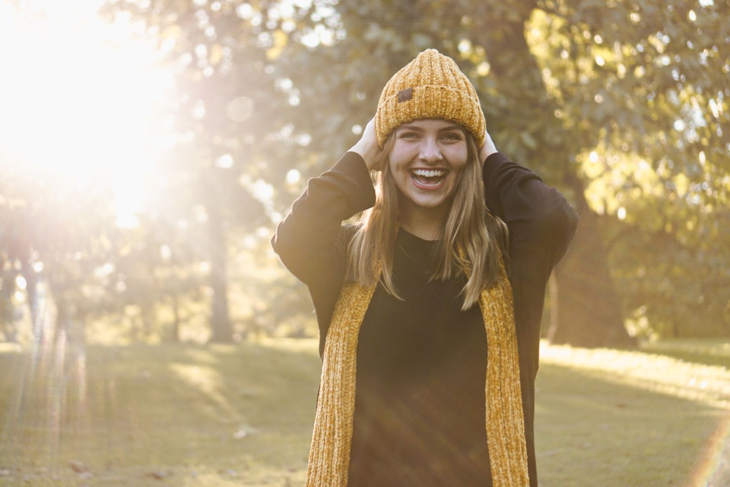 A person in a cozy knit sweater smiles while standing in a sunlit autumn park with golden leaves falling.