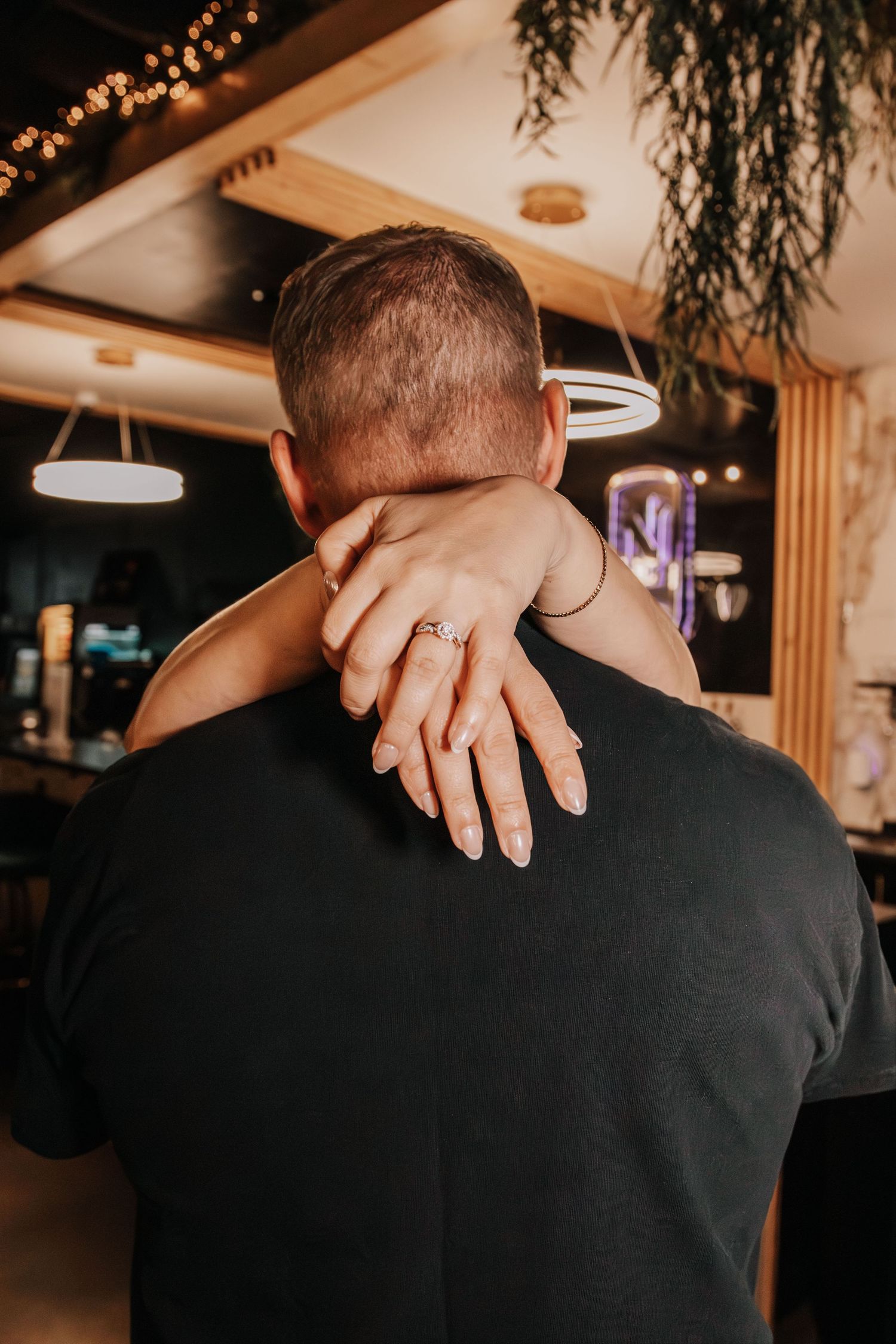 Couple embracing in a restaurant, woman's hand with engagement ring resting on man's shoulder.