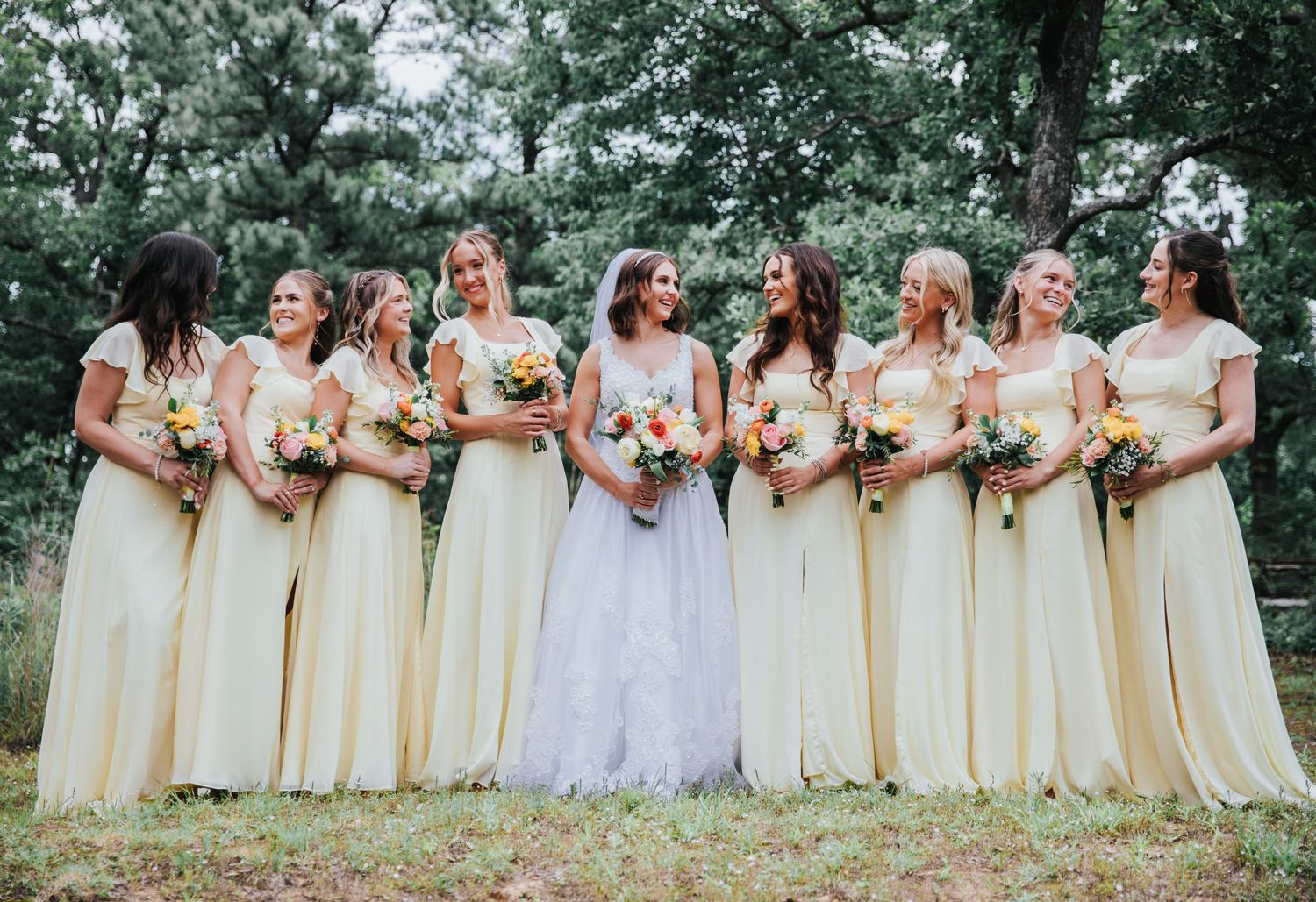 Bride in white lace gown stands with eight bridesmaids in yellow dresses holding floral bouquets outdoors.