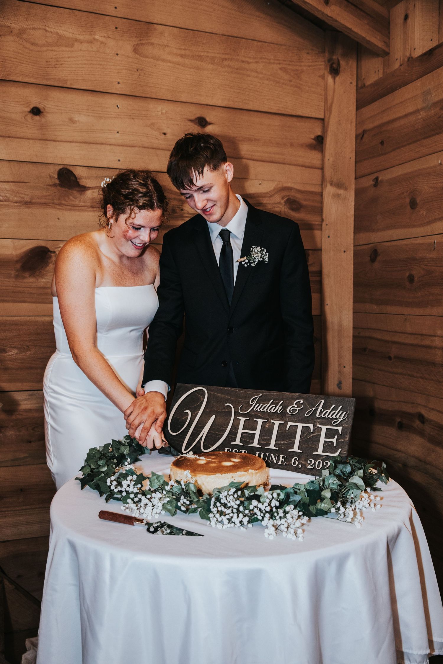 Bride and groom cutting wedding cake at rustic barn reception with White family sign dated June 6.