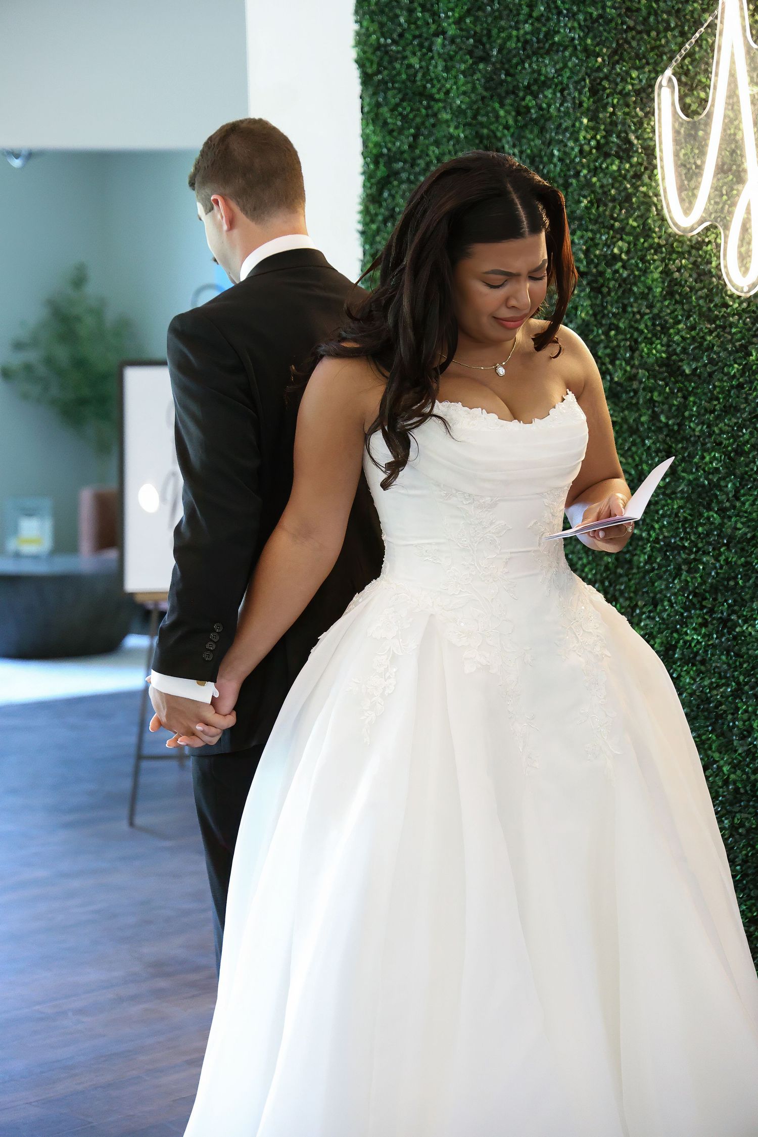 Bride in white ball gown reads vow card while holding hands with groom in black suit during first look ceremony.