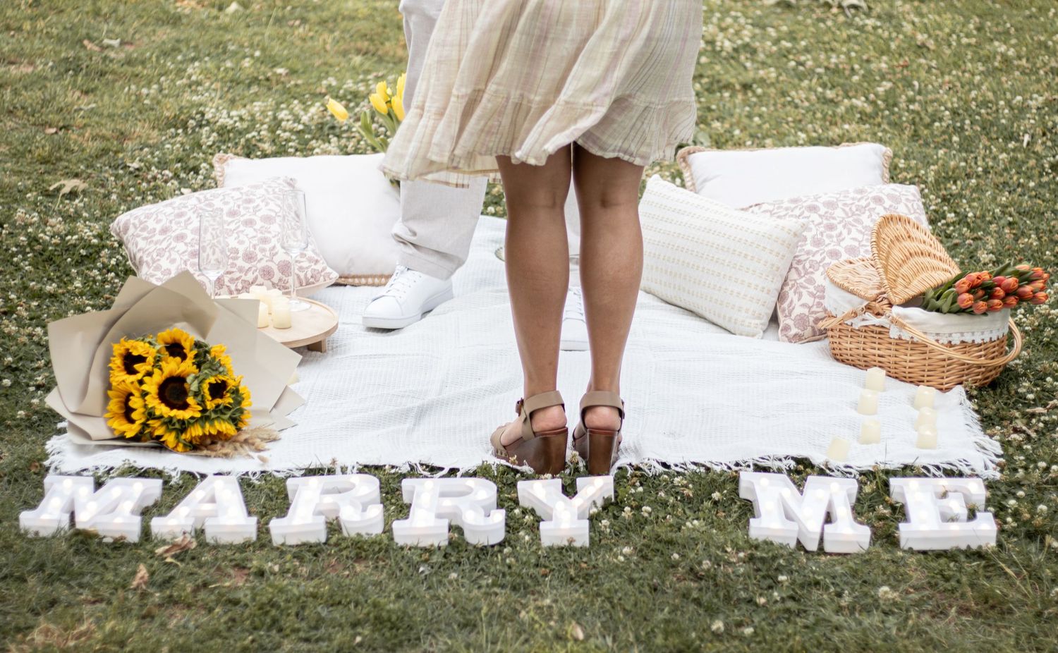 Woman standing on white picnic blanket with sunflowers and 'Marry Me' balloon letters on grass.