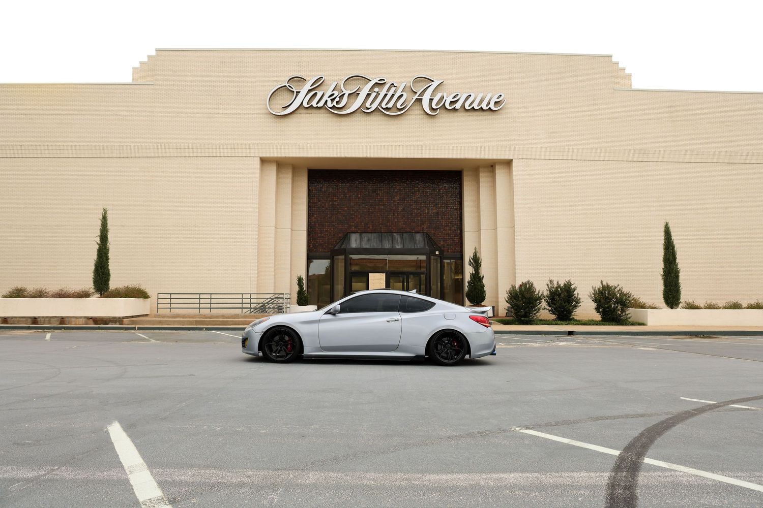 Silver sports coupe parked in front of Saks Fifth Avenue luxury department store in an empty parking lot.